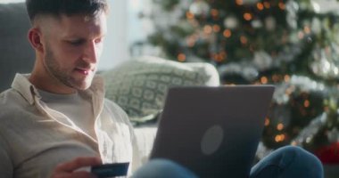 Noel hediyeleri için internetten alışveriş yapan bir adam kredi kartı ve dizüstü bilgisayarını kullanır ve evinde şenlikli bir tatil atmosferiyle çevrilidir..