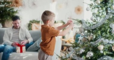 Genç bir çocuk Noel mücevherlerini dikkatlice ağaca asar. Şenlik sezonunun dekorasyonuna yardım ederken yüzü neşe ve konsantrasyonla dolu..