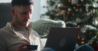 Bir adam internetten kredi kartını ve laptopunu kullanarak güzel dekore edilmiş Noel ağacının önünde, sıcak tatil süslemeleriyle çevrili bir şekilde alışveriş yapar..