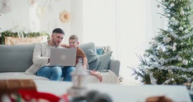 Genç bir çocuk ve babası birlikte oturup internete bakıyorlar ve bir dizüstü bilgisayardan yılbaşı hediyesi alıyorlar. Neşeli ve neşeli bir anın tadını çıkarıyorlar..