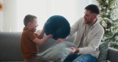 Bir baba ve oğlu, Noel 'de kanepenin üzerinde eğlenceli bir yastık savaşı yapıp neşeli, neşeli ve sıcak aile anlarını neşeli bir atmosferde paylaşıyorlar..