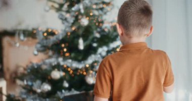 Genç bir çocuk, sıcak, şenlikli oturma odasındaki ağaca parlak bir Noel mücevheri asıyor..