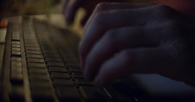 Geceleri bilgisayar klavyesine yazı yazan ellerin yakın plan görüntüsü, karanlık bir odada ekranların yumuşak parıltısıyla aydınlatılıyor..
