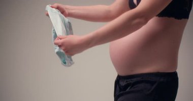 Gelişmiş hamilelik dönemindeki bir kadın, bebeğinin doğmasını heyecanla beklerken, küçük bebek kıyafetlerinin içinden sevgiyle bakar..