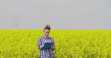 Modaya uygun bir kadın çiftçi arka planda çiçek açan kolza tohumu tarlası olan bir deftere notlar yazıyor. Modern tarım ve tarım belgeleri kavramı.