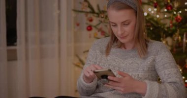 Bir kadın süslenmiş bir Noel ağacının önünde akıllı telefonuyla internetten alışveriş yapıyor.