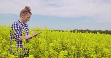 Çiçek açan kolza tohumu tarlasında duran, gülümseyen ve kırsal alanın tadını çıkaran mutlu genç kadın çiftçi. Çiftçilik yaşam tarzı ve ilkbahar tarım kavramı.