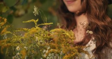 Elinde kır çiçekleri tutan düşünceli genç bir kız ağaçların önünde sessizce duruyor.