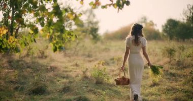 Gün batımında çayırda elinde bir sepet çiçekle mutlu bir kız yürüyor.