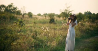 Gülümseyen bir kız açık arazide eski bir kamerayla fotoğraf çekiyor, doğanın ve ışığın tadını çıkarıyor.