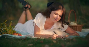 Gülümseyen hayalperest bir kız rüyalarını piknik battaniyesinde yatarken bir deftere yazar.