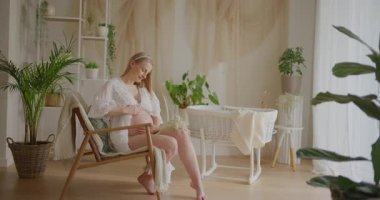 Güzel, hamile bir kadın güzel dekore edilmiş bir odada beşikte poz veriyor, mutlu ve pozitif gülümsüyor..