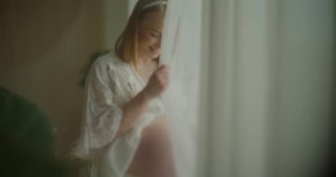 Düşünceli, neşeli hamile bir kadın pencerenin kenarında durur ve çocuklarının doğumunu heyecanla bekler..