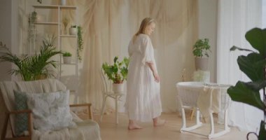 Hamile bir kadın, içinde bebek olan küçük bir beşiğe yaklaşır ve bebeği nazikçe sallamaya başlar..