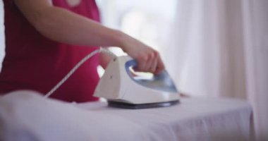 Hamile kadın evdeki ütü masasında yatak ütülüyor. Ev işleri yapmasına rağmen yorgun görünüyor..