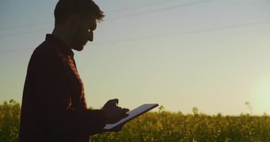 Güzel bir günbatımı ve sarı kolza tohumu tarlasına karşı dijital tablet kullanan bir çiftçinin silüeti modern tarım teknolojisini yakalıyor..