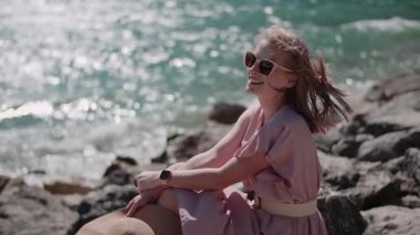 Neşeli bir kadın mavi su ve taş dağlarla kayalık bir sahil kenarında duruyor..