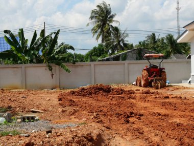 Traktör inşaat alanındaki toprağı beton duvarlı ve arka planda mavi gökyüzü olan ağaçları sürüp düzleştiriyor, araziyi düzleştirmek için makineler kullanıyor.