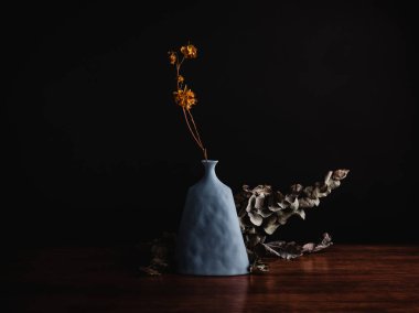 Kil vazoda karamsar bir çiçek, kurumuş yapraklarla birlikte, ahşap bir masa dekorasyonu sanatının karanlık bir odasında izole edilmiş.