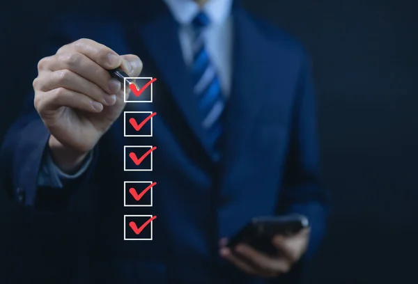 Businessman hand holding a pen ticking a red checkmark on a box in the list Show on the virtual screen. Checklist Ideas in Business Survey items and questionnaires. copy space