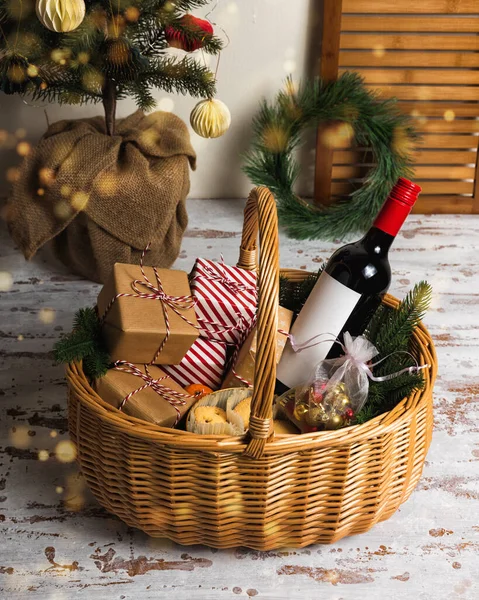 Christmas gift basket with bottle of red wine, gift boxes, cookies, and mandarins. Fir tree with ornaments on rustic table