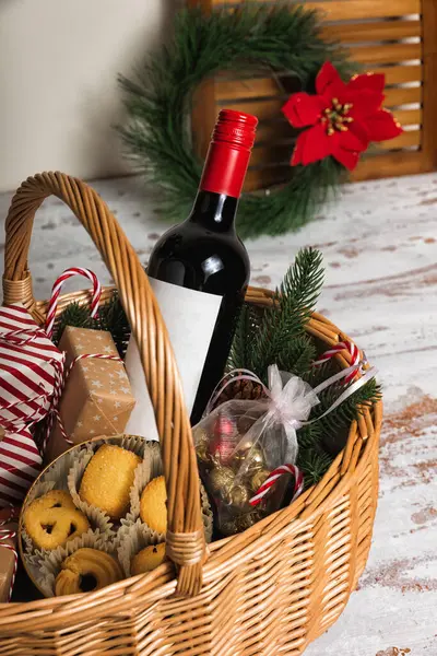 Christmas gift basket with bottle of red wine, gift boxes, cookies, and mandarins. Fir tree with ornaments on rustic table