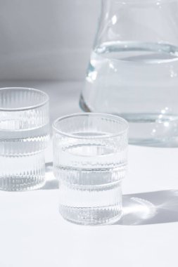 Cam bardaklar ve bir sürahi temiz, temiz içme suyu standı beyaz bir masanın üstünde. Güneşli bir gün ve camdan keskin gölgeler.