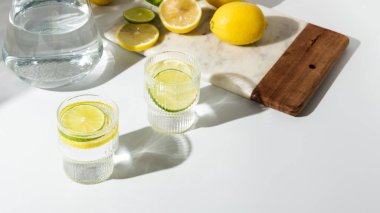 Cam bardaklar ve bir sürahi temiz, berrak limon ve limon suyu beyaz bir masada. Güneşli bir gün ve keskin gölgeler limonata bardağından
