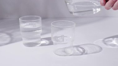 Cam bardaklar ve bir sürahi temiz, temiz içme suyu standı beyaz bir masanın üstünde. Güneşli bir gün ve camdan keskin gölgeler.