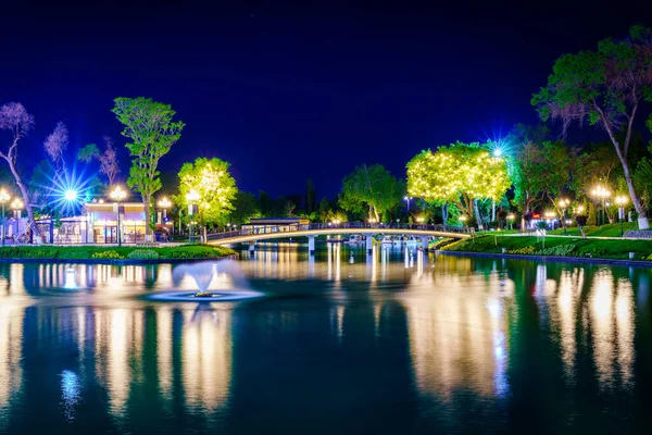 stock image Blurred splashing fountain water flows in an artificial lake in a night park with lanterns, a bridge and trees.