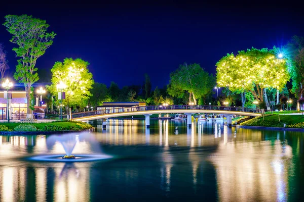 stock image Blurred splashing fountain water flows in an artificial lake in a night park with lanterns, a bridge and trees.