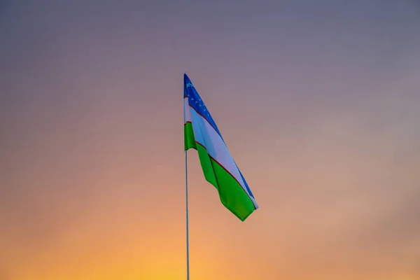 夕日や日の出に揺れるウズベキスタンの国旗劇的な曇りの空の背景 — ストック写真