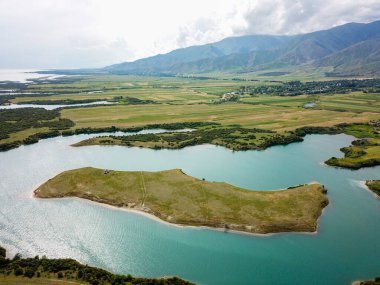 Güneşli bahar günü, arka planda su ve dağlarla çevrili ıssız bir adada otlayan at sürüsü manzarası.