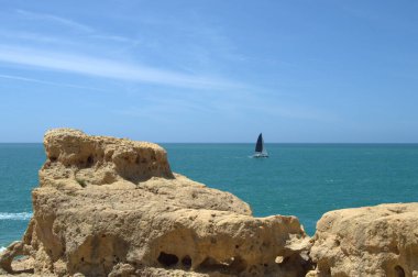 Bir yelkenli, açık mavi gökyüzünün altında, Carvoeiro 'nun kayalık uçurumlarının yanında okyanusu aşar. Engebeli kaya oluşumları dingin deniz manzarasına dramatik bir ön plan ekler.