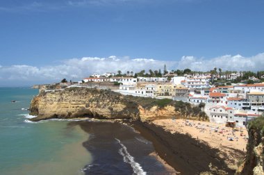 Carvoeiro, Portekiz - 13 Mayıs 2022: Praia de Carvoeiro 'nun güneşli günün keyfini çıkaran plaj, kayalıklar ve turistlerle dolu evleri gösteren panoramik bir manzarası