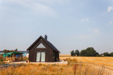Geniş bir pencere ve bacası olan modern siyah kulübe, temiz bir gökyüzünün altındaki geniş altın buğday tarlasında izole edilmiş.