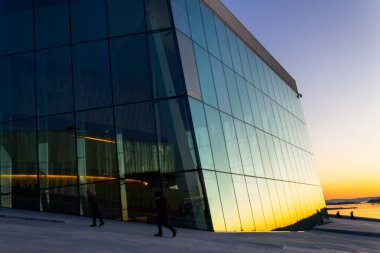 Oslo 'daki Oslo Opera Binası' nın cam duvarı gün batımında şehir manzarasını yansıtıyor.