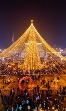 Bishkek, Kırgızistan - 30 Aralık 2023: İnsanlarla dolu bir meydandaki bir Noel ağacının aydınlatma ışıkları havası ve 2024 pankartıyla dönen spot ışıkları