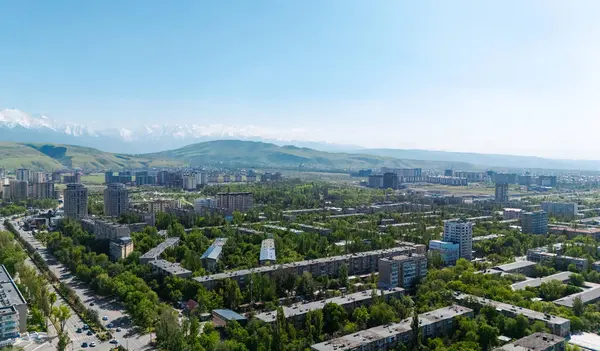 Baharda Bişkek şehrinin hava manzarası, yemyeşil, kentsel binalar ve açık mavi gökyüzünün altında karlı dağlar