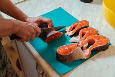 Elinde bıçakla evdeki mavi kesme tahtasına çiğ başsız somon balığı kesiyor.