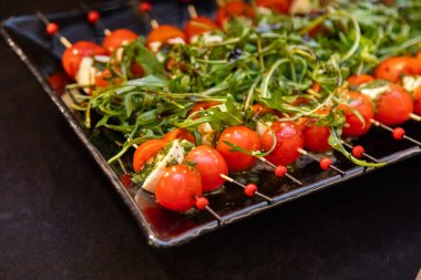 Kirazlı domates ve mozzarella peynirli şiş yakın plan, siyah tepside balzamik jöle serpiştirilmiş taze roka ile zarif bir şekilde eşleştirilmiş.