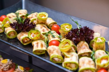 İştah açıcı kabak dürümleri lezzetli bir dolma ile doldurulmuş, taze domates ve yeşilliklerle süslenmiş, parlak siyah bir tepside sergilenmiş.