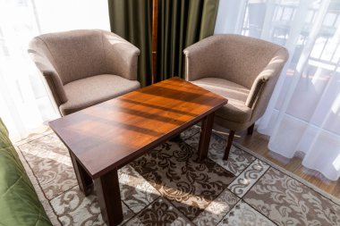 Modern hotel room featuring two upholstered chairs and a polished wooden table, set against curtains and a bright window clipart