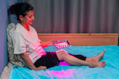 Senior woman treating sore leg with the red light therapy handheld device at home on the bed clipart