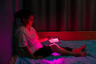 Senior woman treating sore leg with the red light therapy handheld device at home on the bed clipart