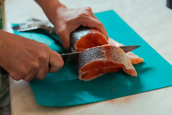 stock image Hand with a knife cutting raw headless salmon fish laying on blue cutting board at home