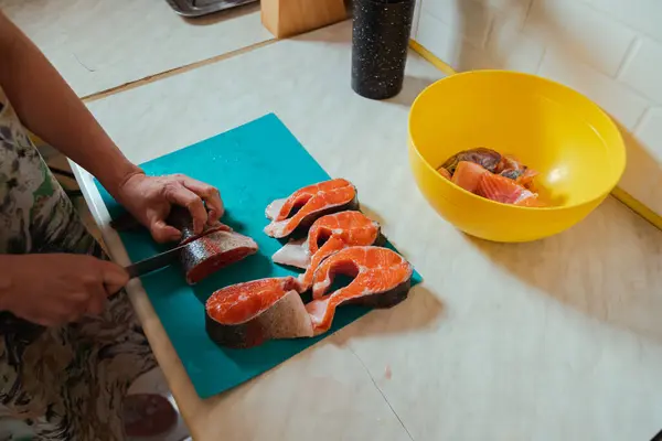 stock image Hand with a knife cutting raw headless salmon fish laying on blue cutting board at home