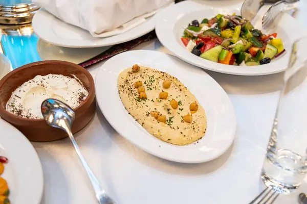 stock image A spread of Mediterranean appetizers including hummus topped with chickpeas, Greek salad, and whipped cream dip in a brown clay bowl on a white dining table