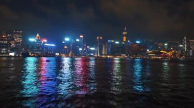 Limanın karşısından Hong Kong 'un gökdelenlerinin ve şehrin ışıltılı ışıklarının suya yansıyarak çarpıcı bir şehir manzarası oluşturduğu gece manzarası.