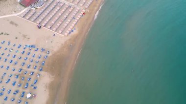 Plaj şemsiyeleri ve şezlonglarıyla kumlu sahilin aşağı havadan görünüşü.